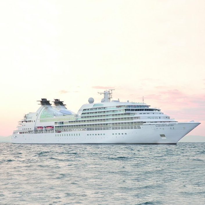 Cruise ship sails on calm ocean during sunset, casting reflections with pink-tinged sky.