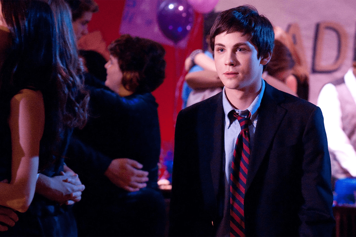 Young man stands wearing a suit and striped tie, surrounded by dancing people at a dimly lit party with balloons.