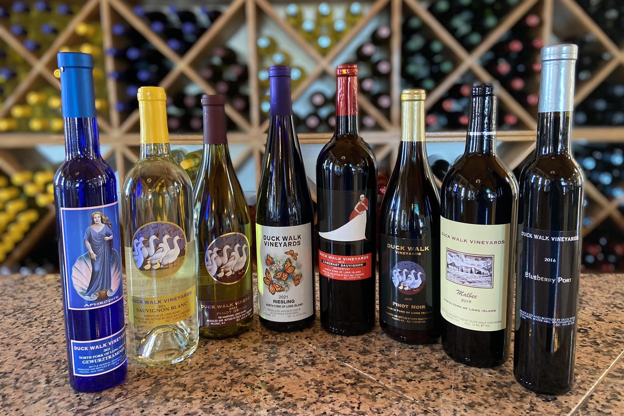 Bottles stand in a row displaying Duck Walk Vineyards wine labels, set on a granite counter, with a grid of colorful wine bottles behind.
