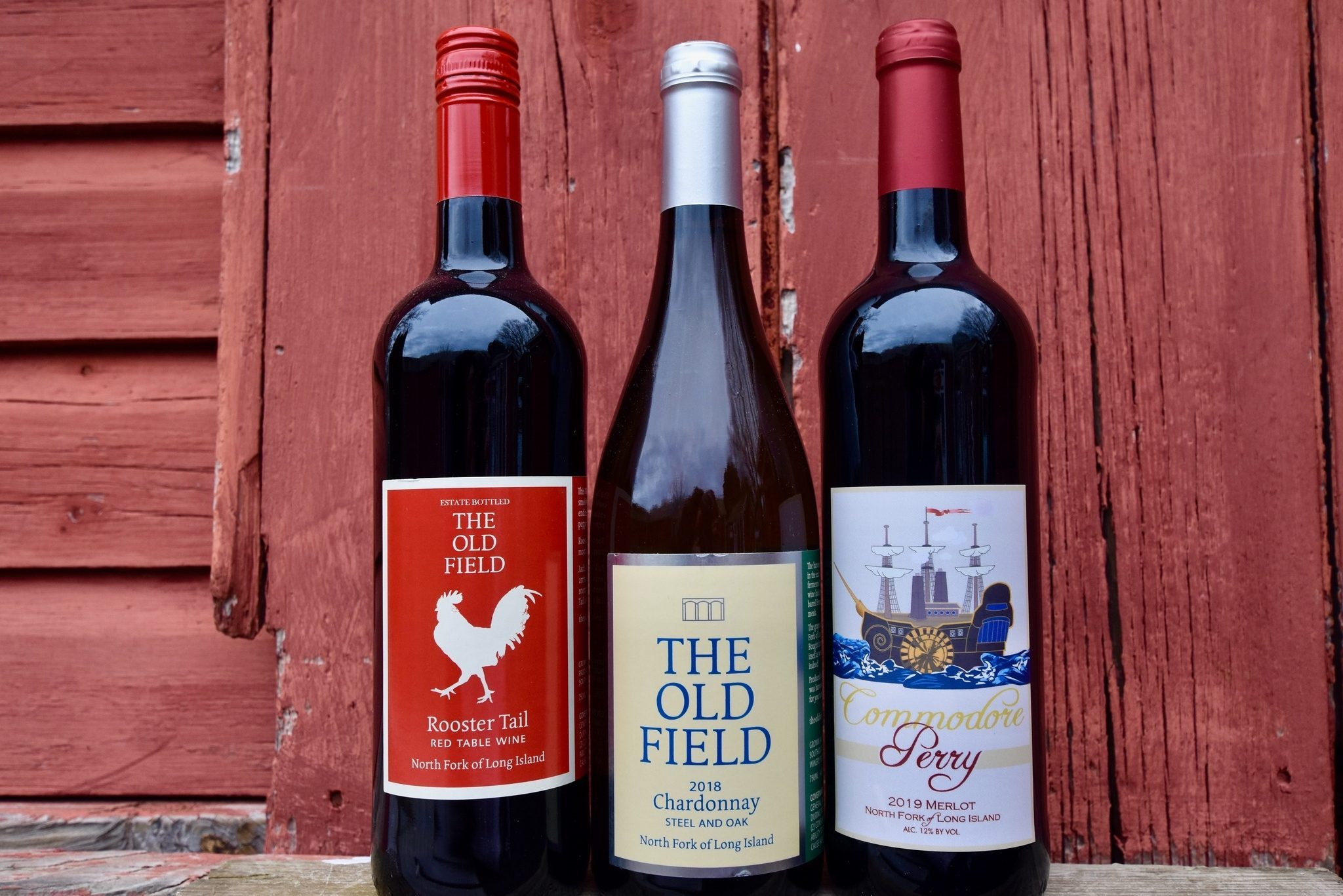 Three wine bottles stand upright against a rustic wooden wall. Labels indicate red and white varieties from North Fork of Long Island.