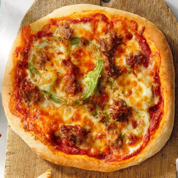 Pizza rests on a wooden board, topped with melted cheese, green peppers, and browned sausage in a warm kitchen setting.
