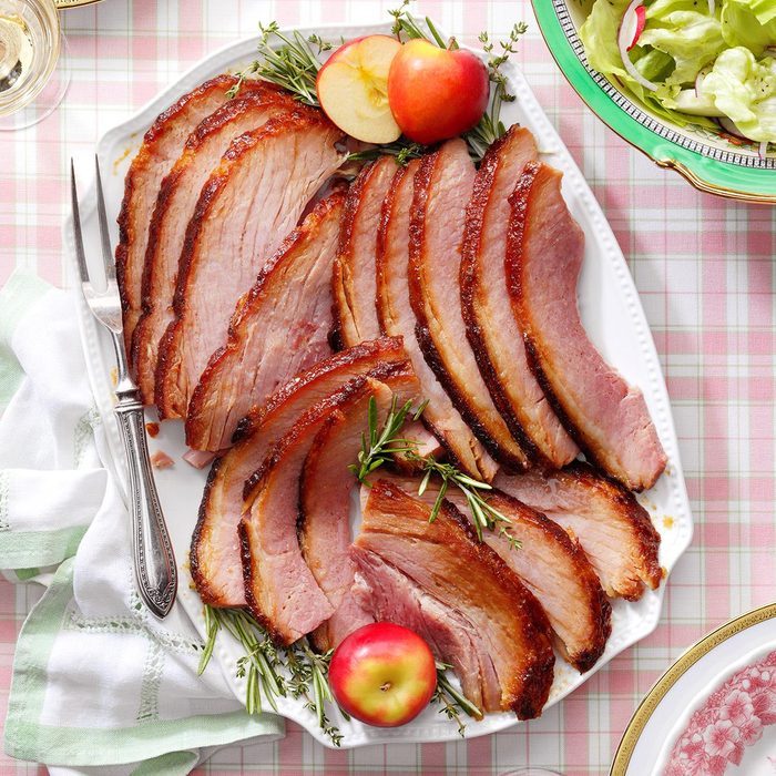Slices of glazed ham, arranged on a white platter, garnished with apples and herbs, set on a pink-checkered tablecloth alongside a salad.
