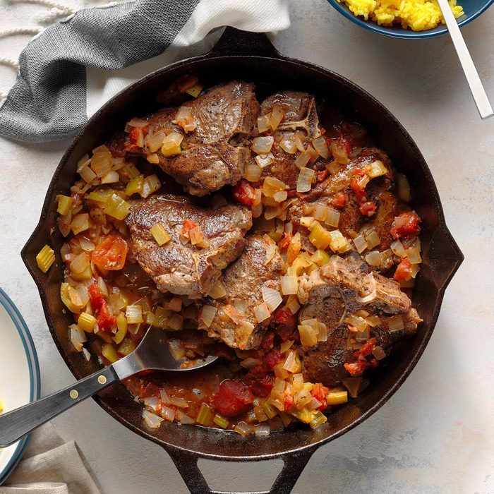 Lamb chops simmering with diced onions, tomatoes, and herbs in a black skillet; a striped cloth and yellow rice bowl nearby on the table.