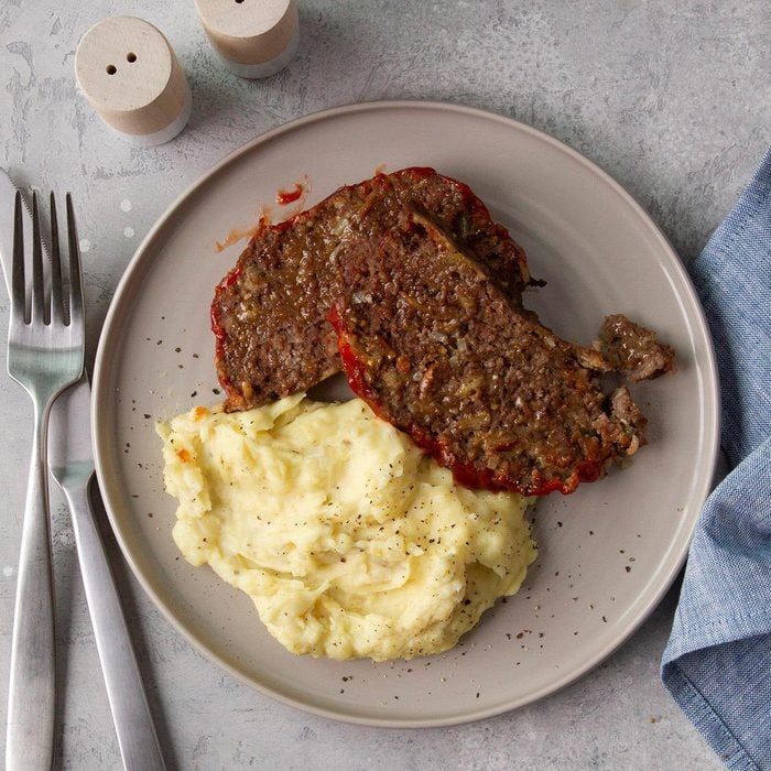 A plate holds mashed potatoes and meatloaf slices, accompanied by cutlery and salt shakers. A blue cloth lies nearby on the table.