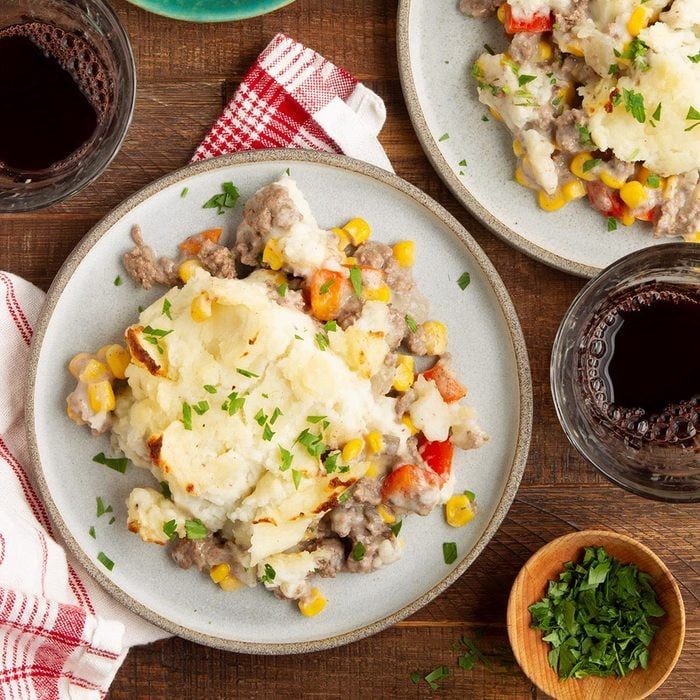 Plates hold shepherd's pie with mashed potatoes and vegetables, accompanied by glasses of red wine and a small bowl of chopped parsley on wooden table.