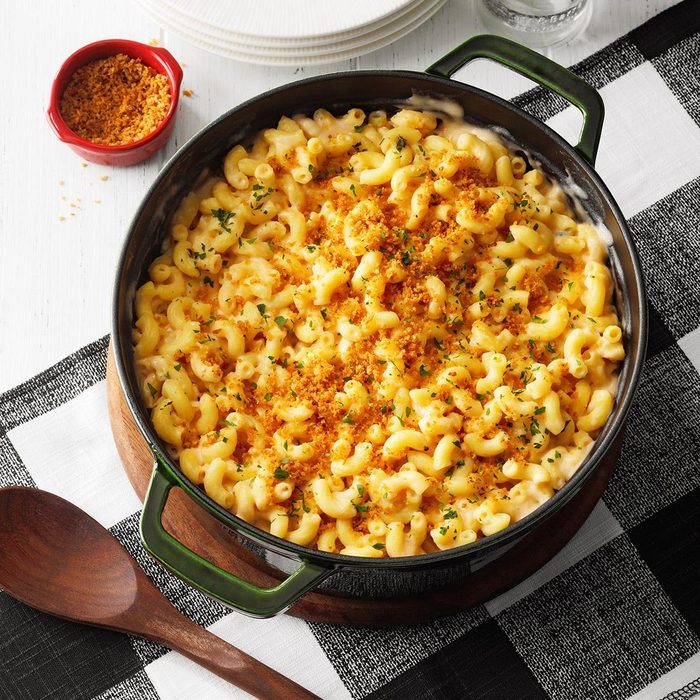 Macaroni sits baked in a green pot, topped with breadcrumbs and herbs, on a checkered tablecloth with a wooden spoon nearby.