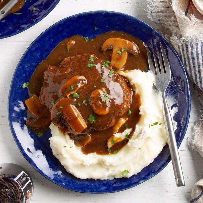 Steak covered in mushroom gravy sits atop mashed potatoes on a blue plate, surrounded by a fork and rustic napkin on a light wooden table.