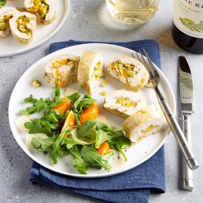 Sliced stuffed chicken rests on a white plate, accompanied by a fresh arugula salad with vibrant tomatoes, set on a gray, textured surface.