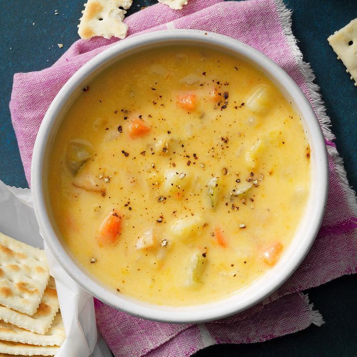 Bowl of creamy vegetable soup sits on pink cloth, with scattered crackers nearby on a blue surface.