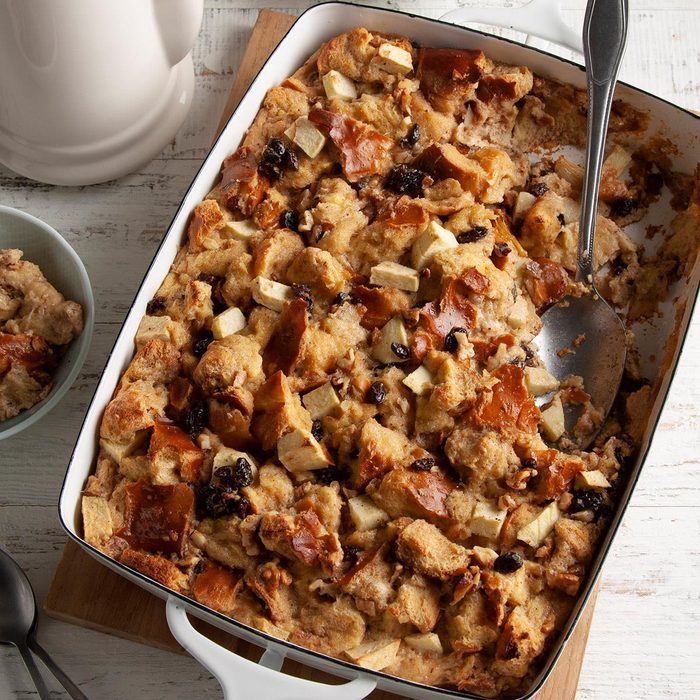 Bread pudding rests in a rectangular dish, scooped partially, with a spoon, on a wooden table, near a jug and bowl.
