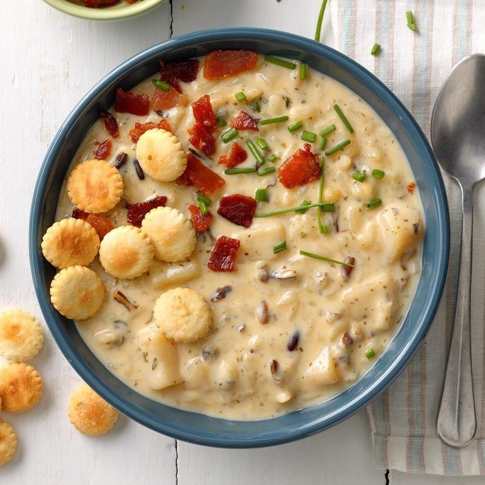 Soup with crackers, bacon, and chives sits in a blue bowl on a striped napkin, next to a spoon and white table.