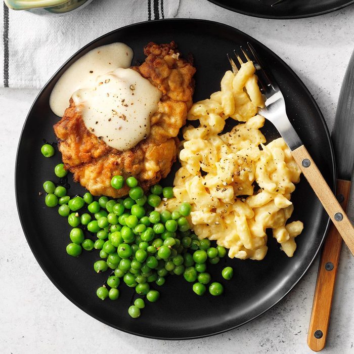 A black plate holds fried chicken with gravy, creamy macaroni and cheese, and green peas, set on a white table with fork and knife.