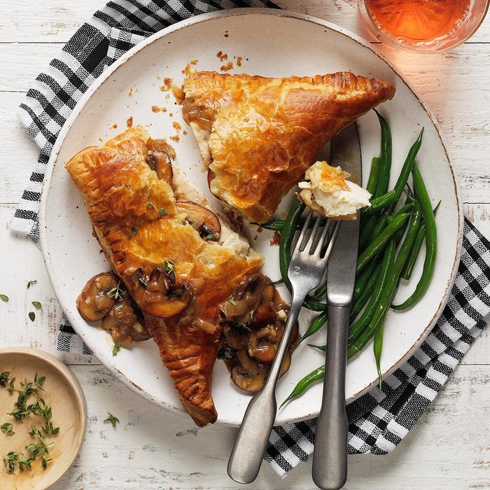 Pastries filled with mushrooms rest on a white plate beside green beans and cutlery, set on a checkered cloth with a drink nearby.
