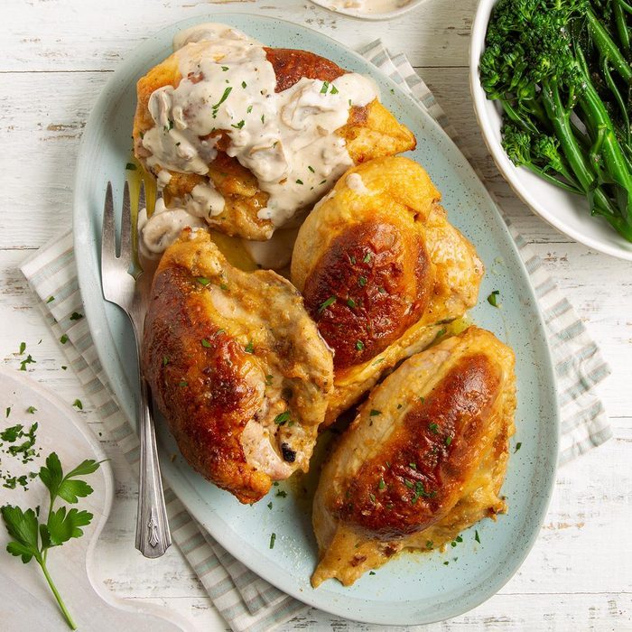 Chicken breast sits on a plate, covered in creamy sauce, alongside a fork, with a side of vibrant green vegetables on a wooden table.