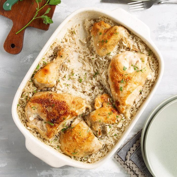 Roasted chicken pieces rest on a bed of seasoned rice in a white baking dish, accompanied by parsley on a cutting board and plates nearby.