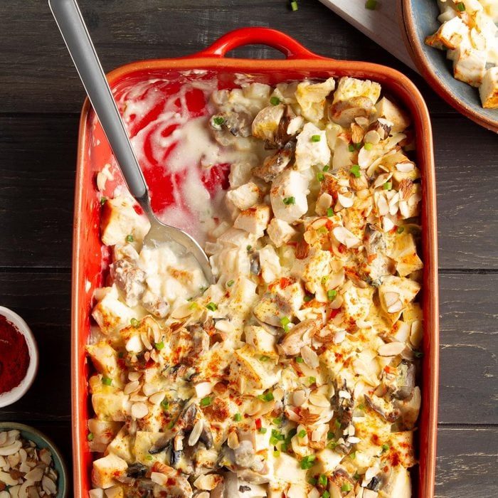 Casserole dish holds creamy, baked chicken and mushroom casserole with sliced almonds, served on a dark wooden table with a metal serving spoon.