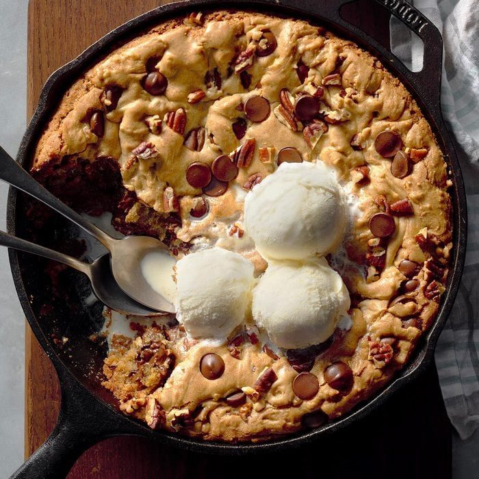 Skillet cookie topped with melting ice cream scoops, surrounded by chocolate chips and pecans on a wooden board.