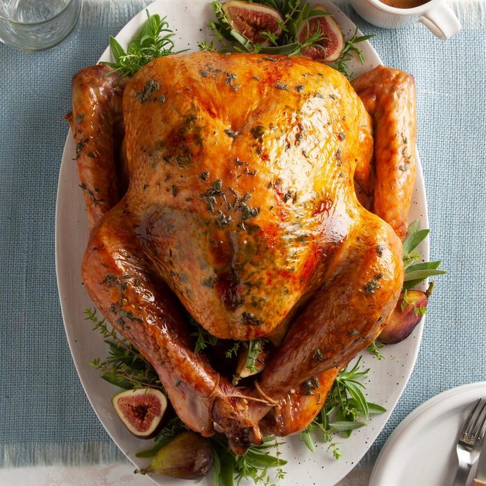 Roasted turkey sits on a platter, garnished with herbs and figs, surrounded by dishes on a blue tablecloth.