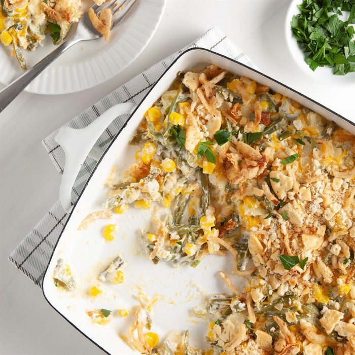 Casserole with cheesy vegetables bakes in white dish on a checkered towel, with a portion served on a plate beside chopped parsley in a bowl.