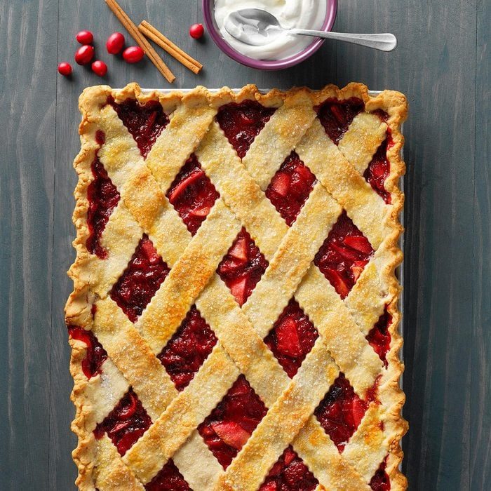 A lattice-topped pie rests on a table, surrounded by cinnamon sticks, red berries, and a bowl of whipped cream with a spoon.