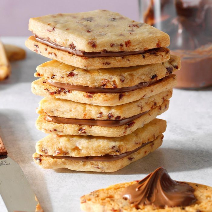 Cookies stacked, filled with chocolate spread, sit on a light surface next to a jar and knife with spread in the background.