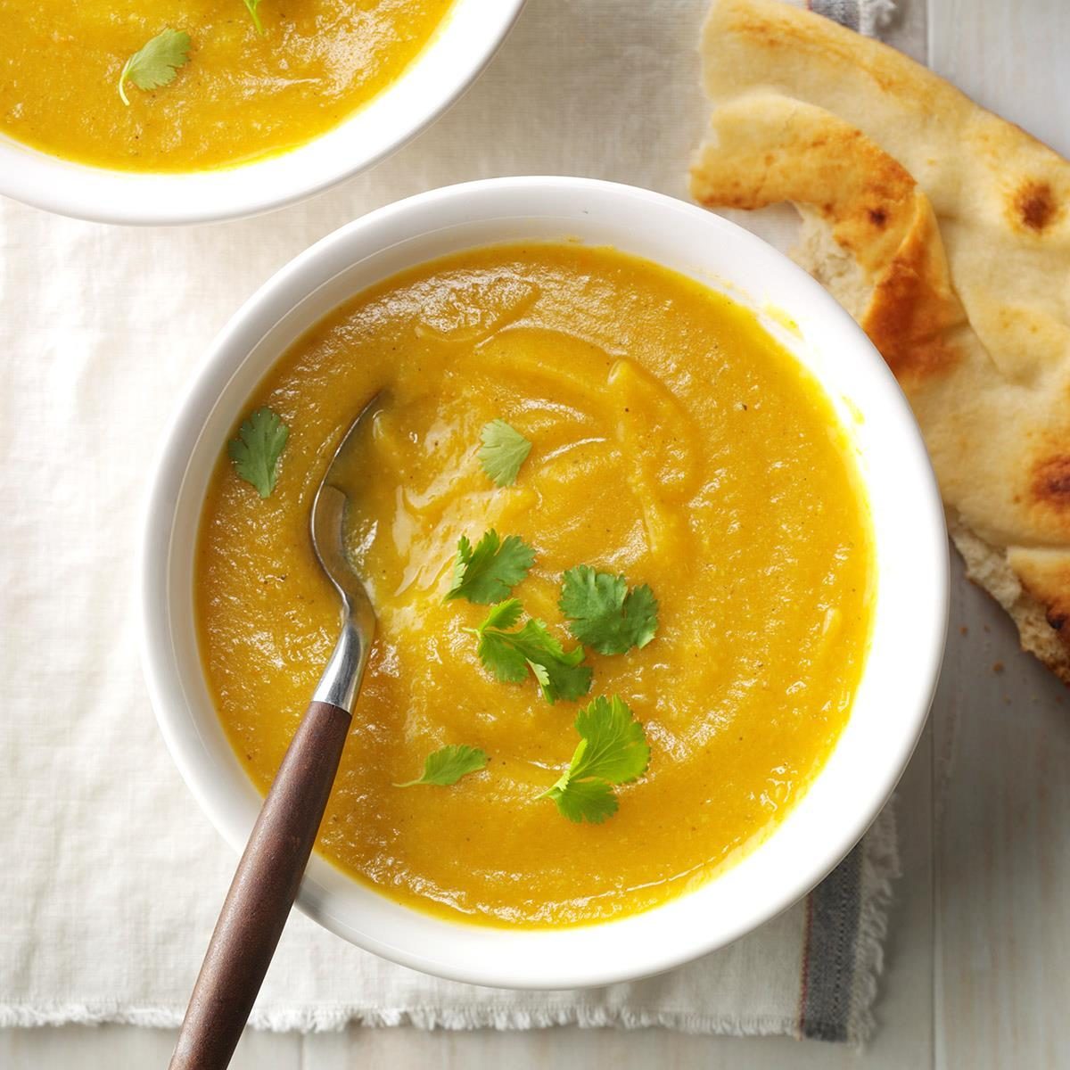 Soup sits in a bowl, garnished with cilantro, accompanied by a spoon and naan bread on a light tablecloth.