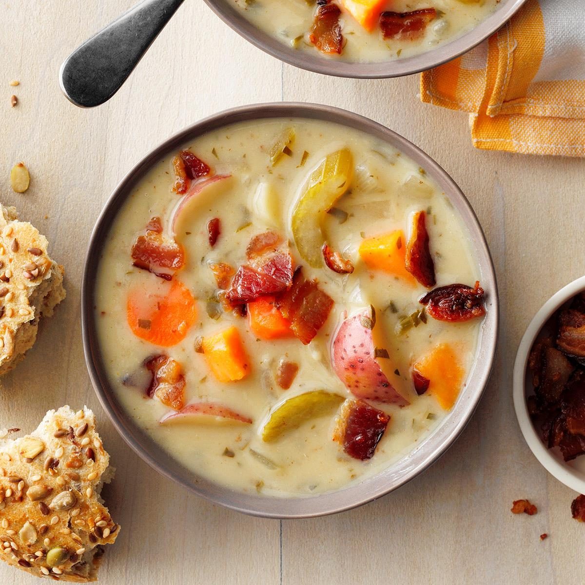 Soup rests in a bowl, featuring bacon, potatoes, and vegetables, with a side of seeded bread on a wooden table.