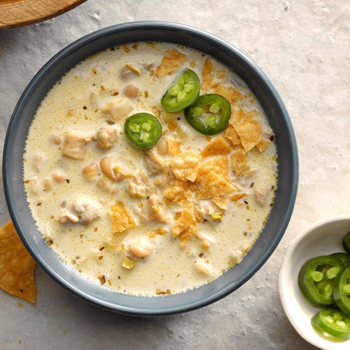 Soup holds ingredients including chicken, beans, and tortilla chips, garnished with jalapeÃ±o slices; served in a gray bowl on a light surface.