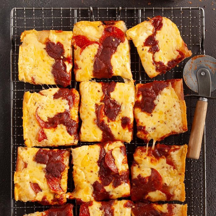 Rectangular pizza slices rest on cooling rack; cheese and pepperoni visible. Pizza cutter with wooden handle lies beside in a kitchen setting.