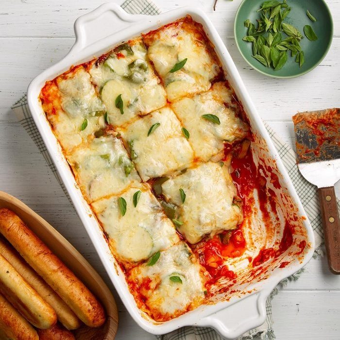 Cheesy lasagna sliced in a white baking dish with one piece missing, surrounded by breadsticks and a bowl of fresh herbs on a white table.