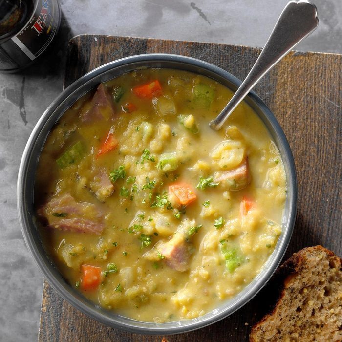 A bowl of chunky split pea soup containing ham, carrots, and celery sits on a wooden surface, accompanied by a slice of bread.