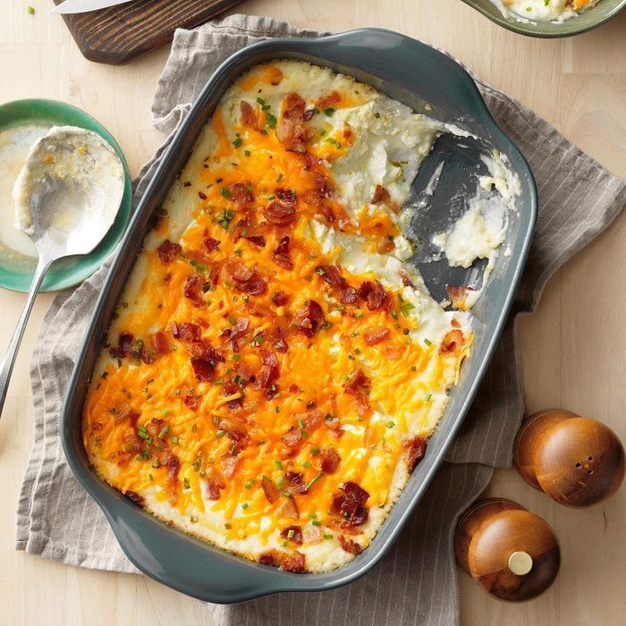 Casserole dish holds mashed potatoes topped with melted cheese and bacon, partially served; on wooden table with a spoon, plate, and salt shakers nearby.