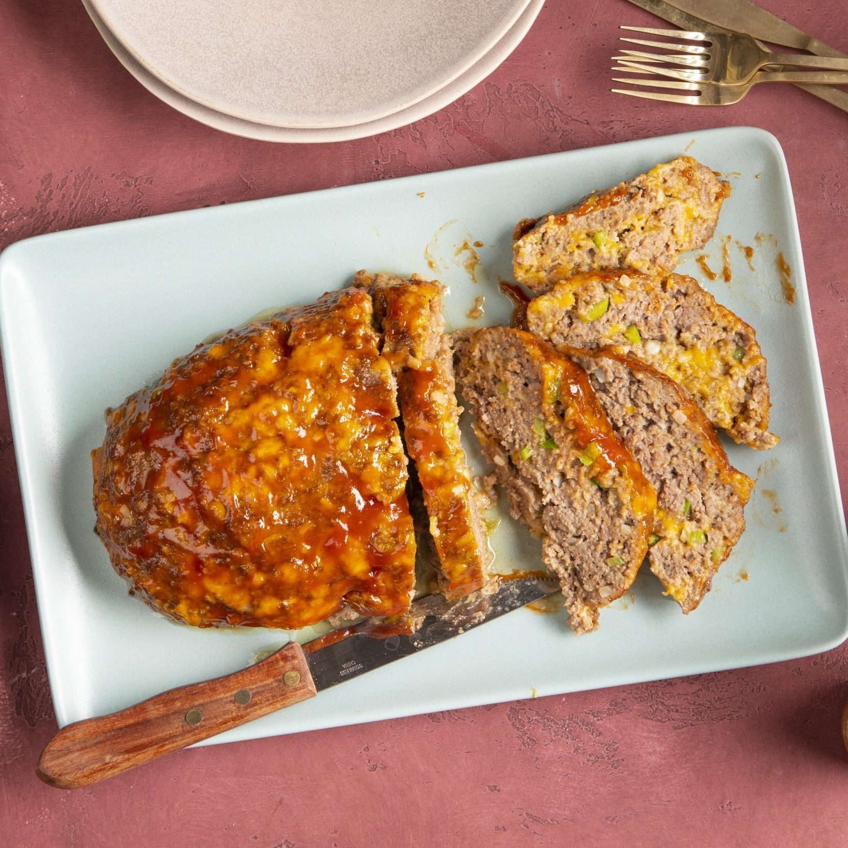 Meatloaf sits on a rectangular plate, partially sliced with a knife; accompanied by empty plates and utensils on a red surface.