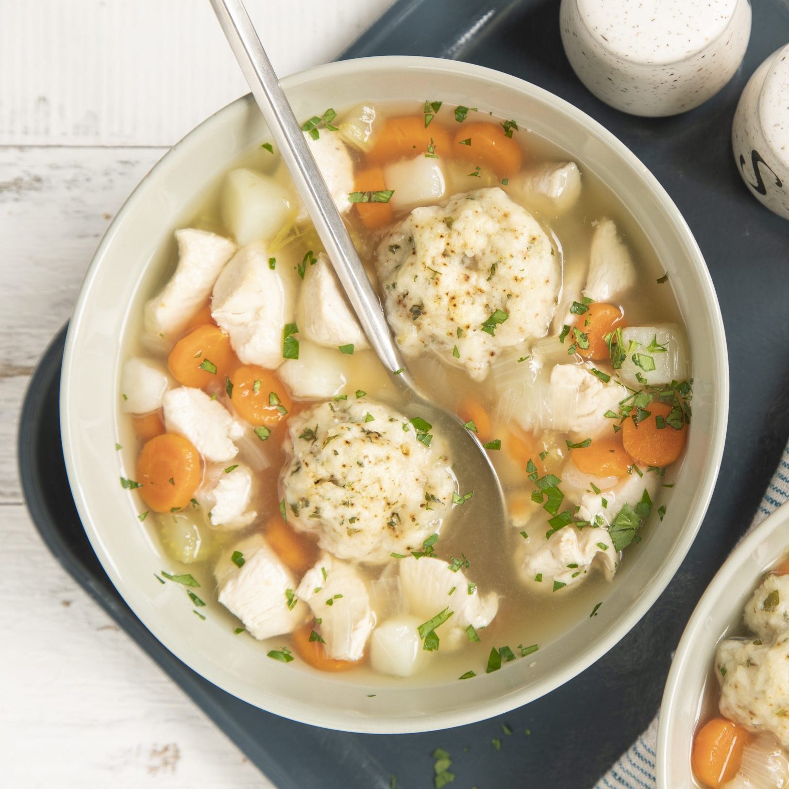 Soup bowl holds dumplings, chicken, carrots, garnished with herbs; spoon rests inside, placed on a dark tray with a salt and pepper shaker nearby.
