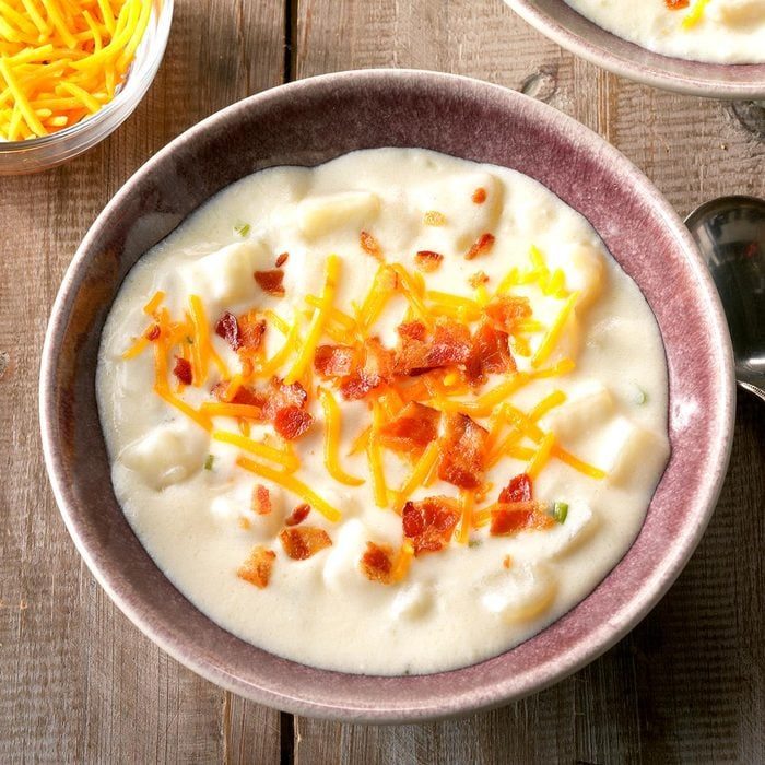 Creamy soup topped with bacon and cheese fills a bowl on a wooden table, with extra shredded cheese in a nearby bowl.