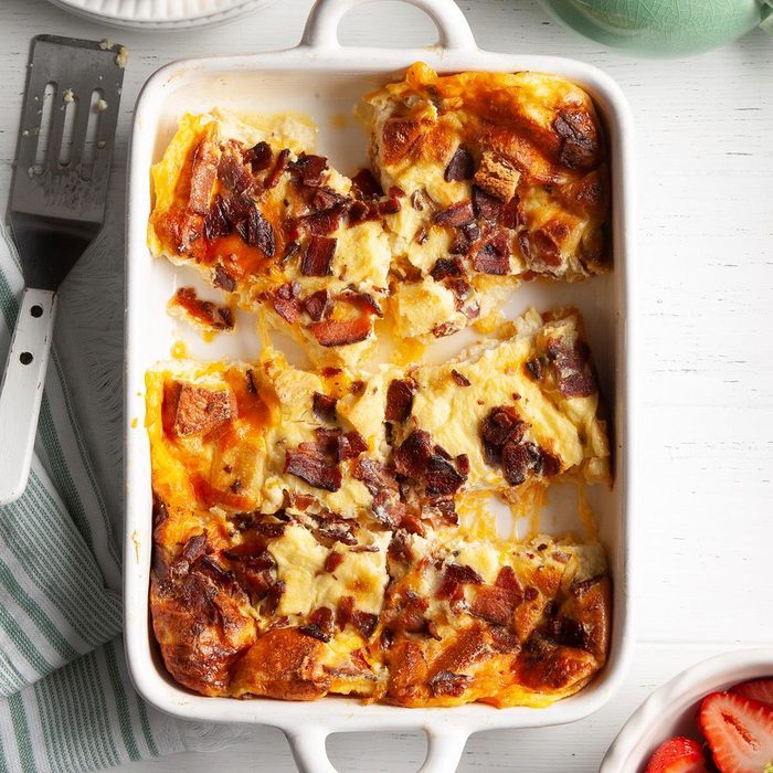 Casserole with melted cheese and bacon sits partially served in a white dish, placed on a white tabletop with a nearby spatula and striped cloth.