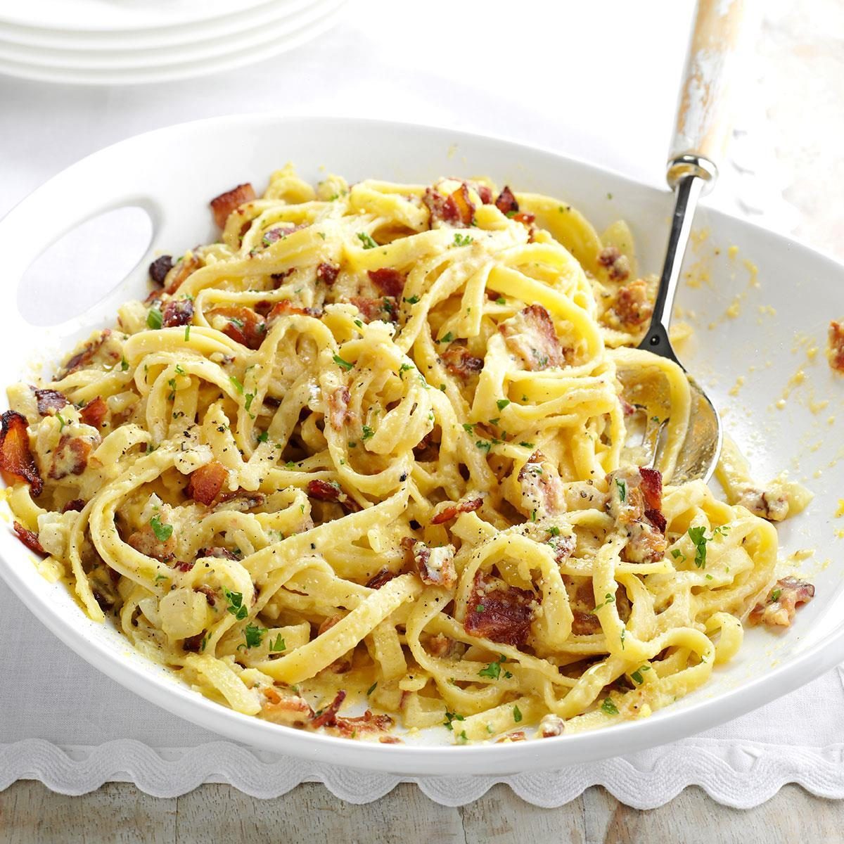 Pasta sits in a white bowl, mixed with a creamy sauce and bacon bits, garnished with chopped herbs on a wooden table.