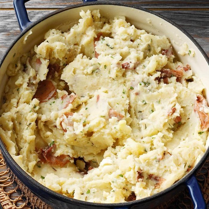 Mashed potatoes rest in a blue pot, garnished with herbs and bits of potato skin, atop a woven placemat on a wooden surface.