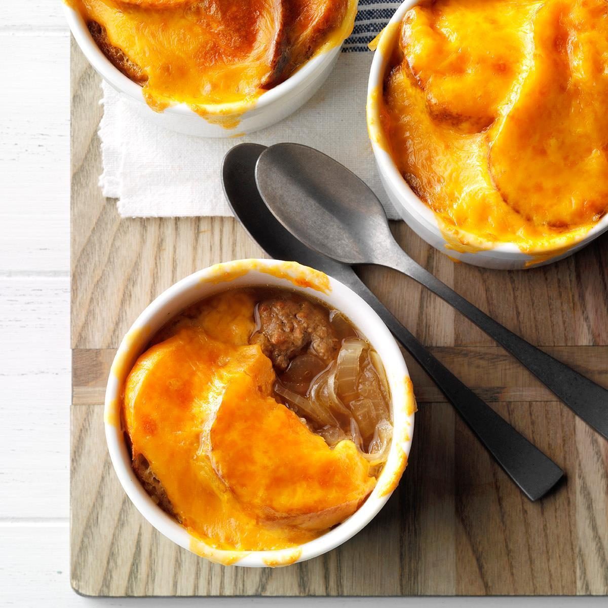 Ramekins filled with baked cheese-topped onion soup, placed on a wooden board beside two metal spoons and a napkin.