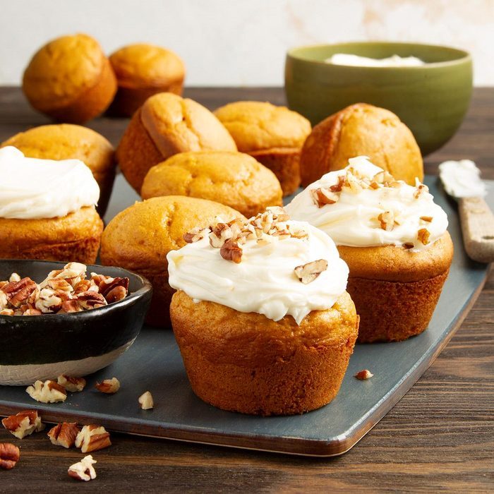 Cupcakes topped with cream and nuts sit on a tray, surrounded by more plain cupcakes and bowls of nuts in a kitchen setting.
