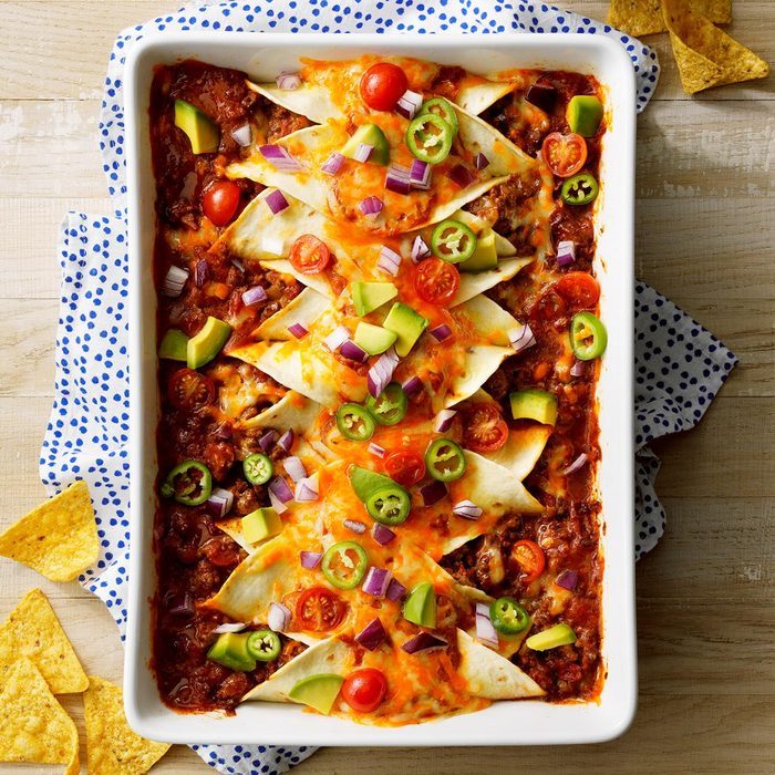 Casserole dish holds layered tortillas topped with melted cheese, cherry tomatoes, avocado, jalapeÃ±os, and onions, on wooden table with tortilla chips nearby.