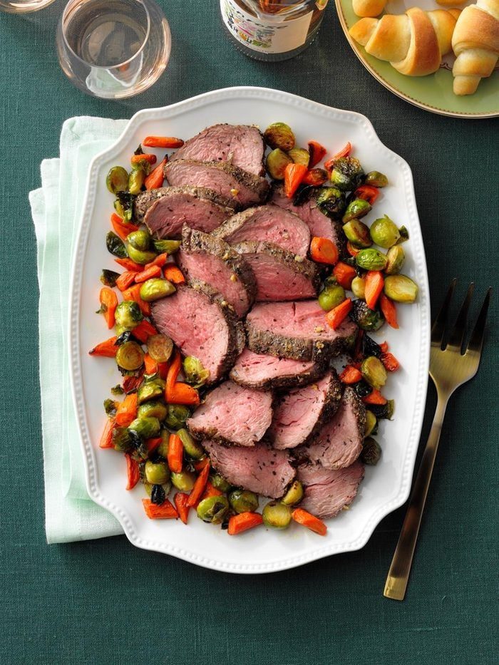 Sliced beef surrounded by roasted vegetables sits on a white platter, atop a green tablecloth next to a glass of wine and croissants.