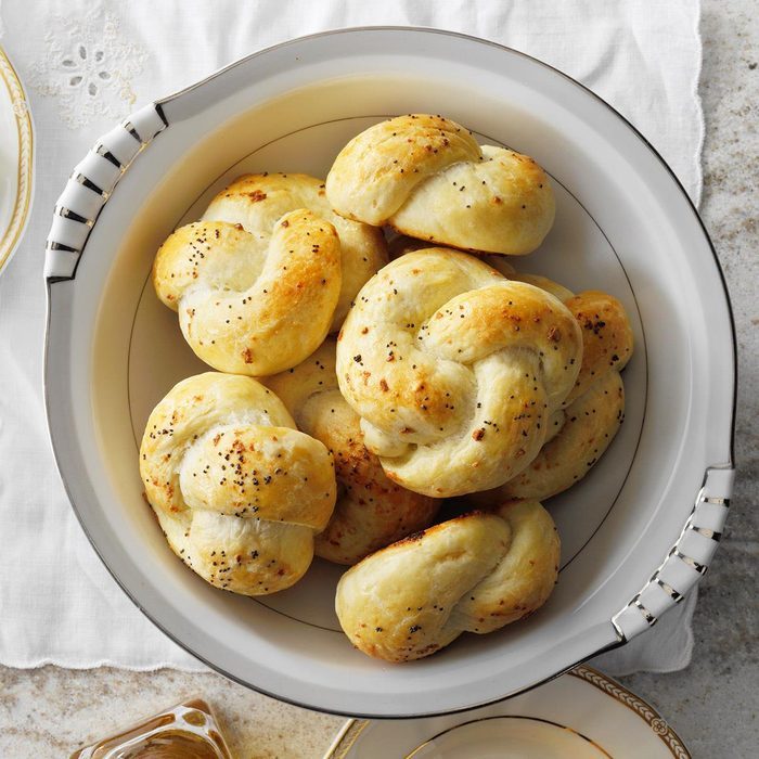 Knotted rolls rest in a white bowl on a table, sprinkled with herbs, suggesting a freshly baked presentation at a dining setting.