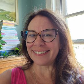 A woman smiles in a room, wearing glasses and a pink top, with a stack of books and a painting in the background.