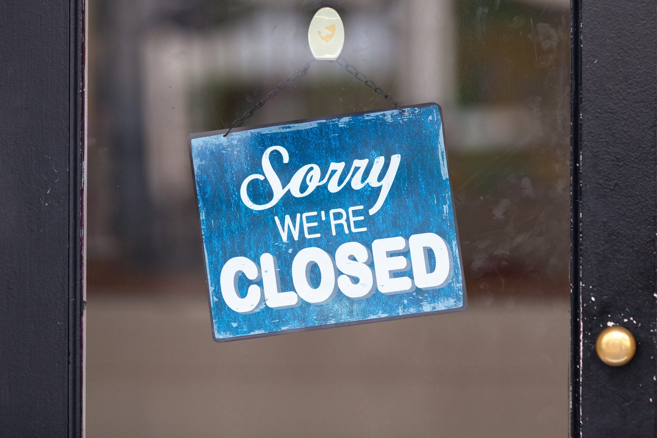 A blue sign reads "Sorry we're CLOSED," hanging on a glass door, set against a blurred indoor background.