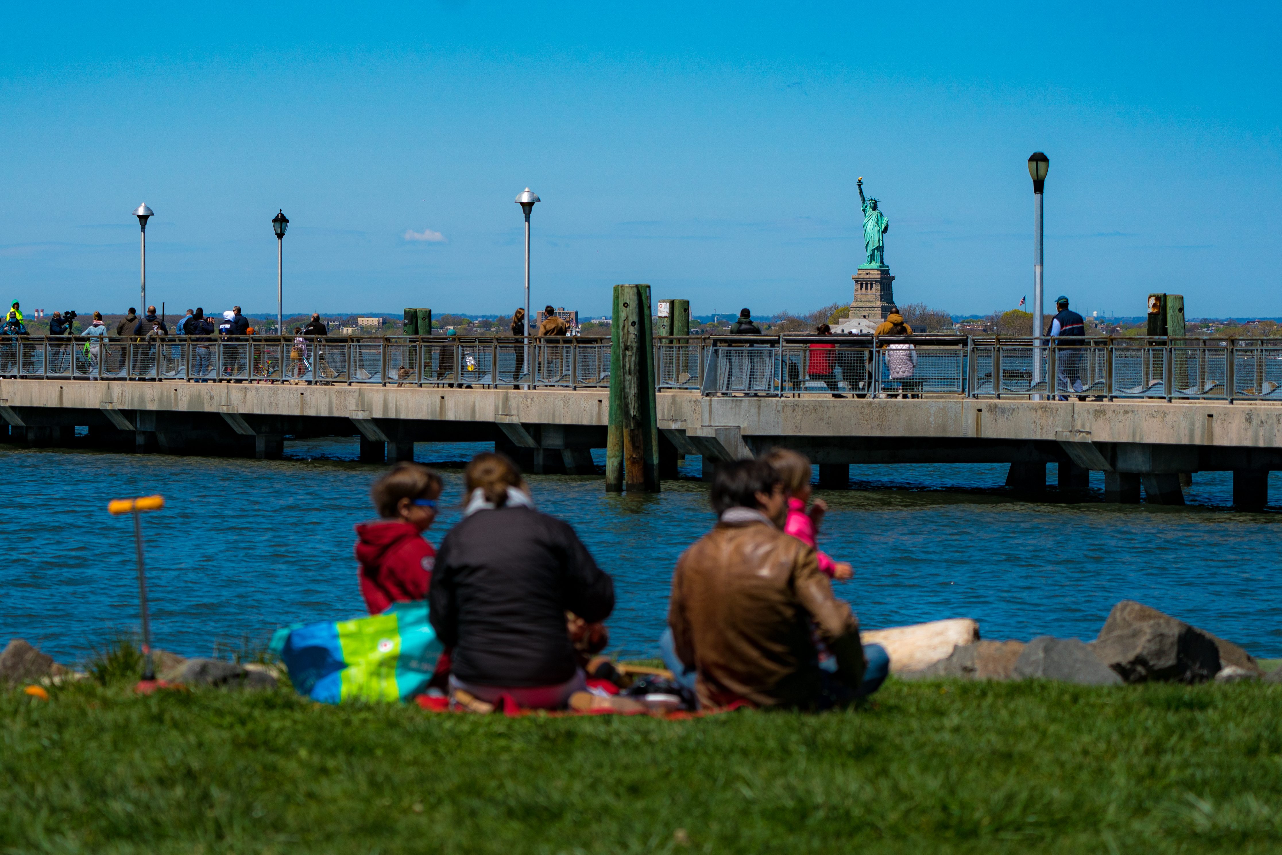 People sit on grass by water, watching a pier with pedestrians. The Statue of Liberty is visible across the water in the background.