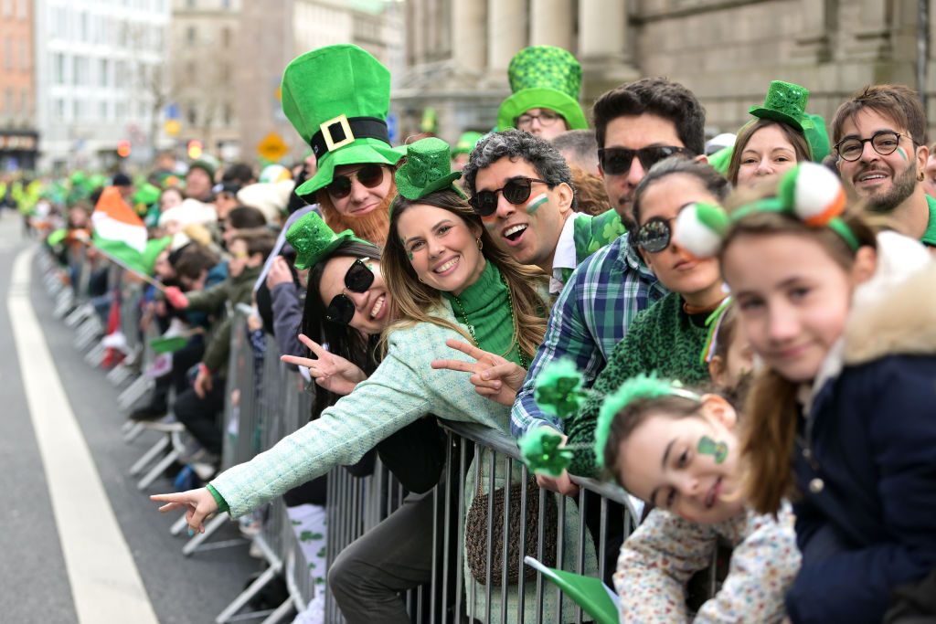 People in festive green outfits cheer behind barriers during a parade, with buildings in the background.