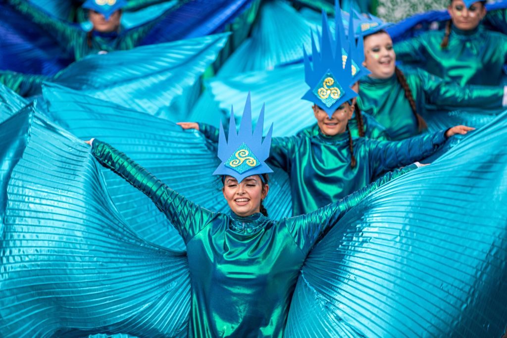 Performers wear blue costumes with pleated wing-like extensions, smiling and dancing in formation. Bright headdresses feature decorative patterns, creating a vibrant, festive atmosphere.