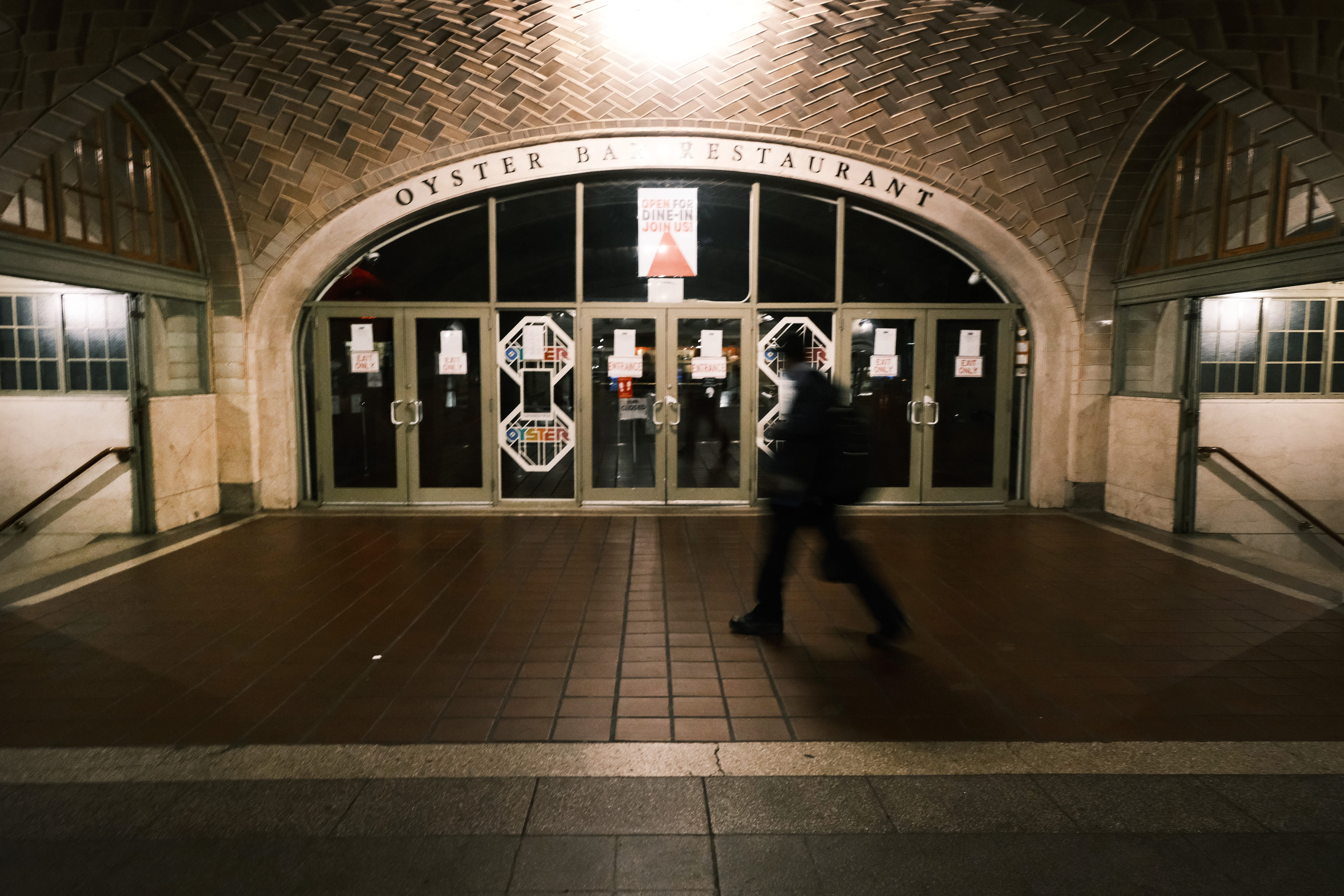 Person walking past closed Oyster Bar Restaurant doors, tiling overhead, in dimly lit hallway. Signs include "OPEN FOR DINE IN. JOIN US!"