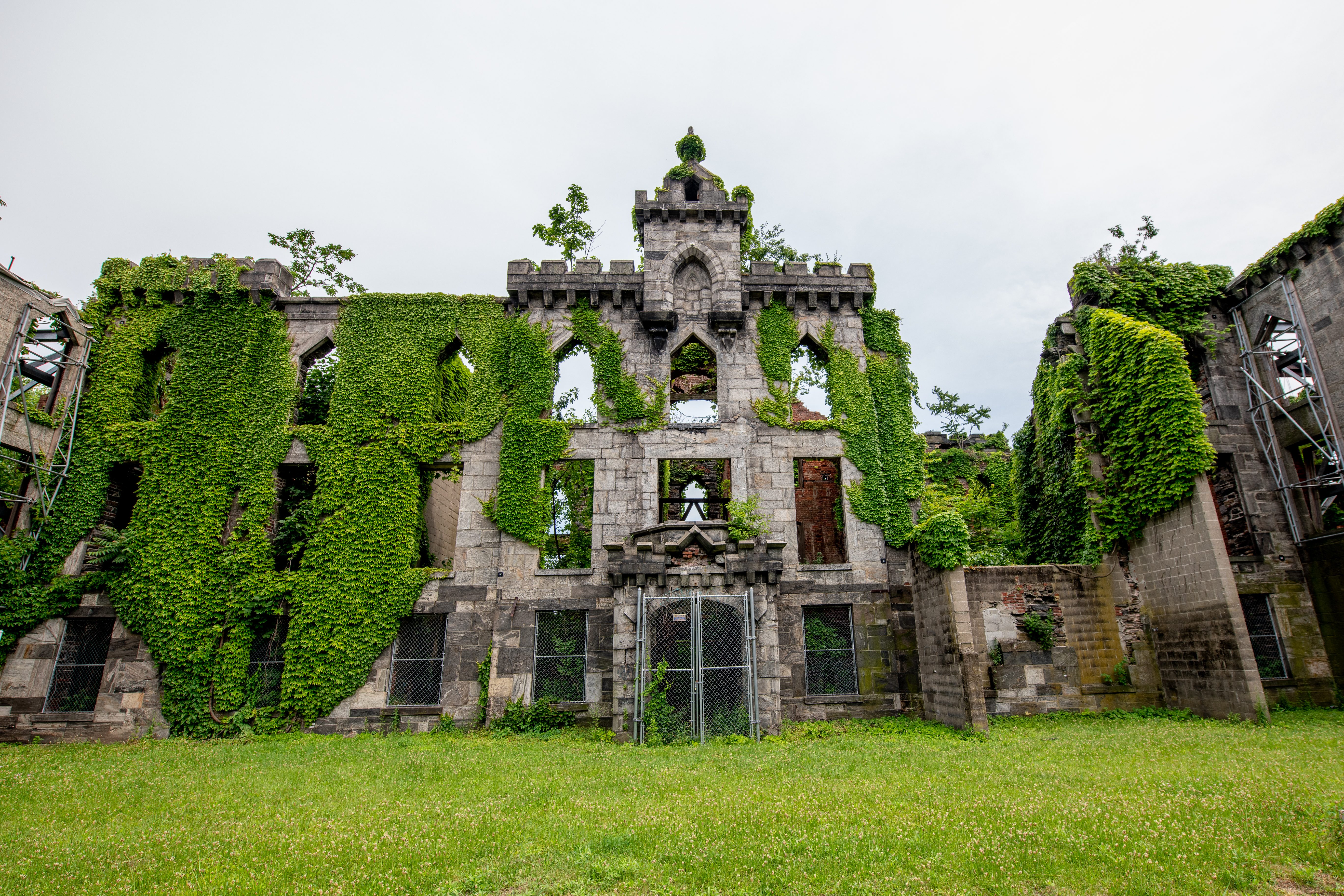 Ruined stone building covered in ivy stands surrounded by grass under a cloudy sky, with scaffolding visible on the side.